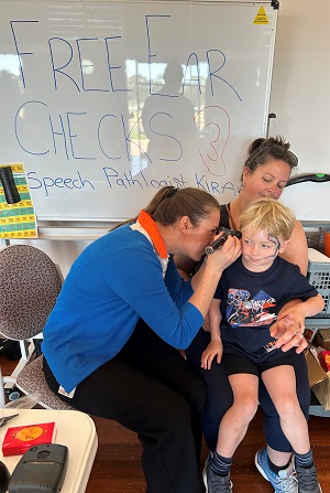 Photo of two adult women with a young toddler. One of the woman is doing a hearing check on the toddlers right ear