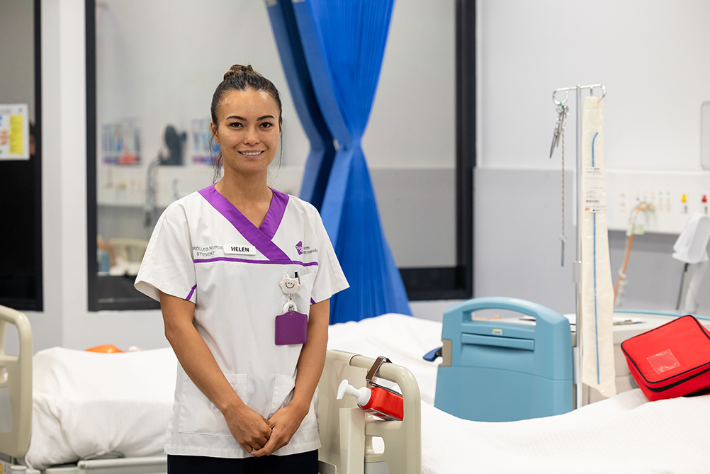 A female nursing student smiling in a hospital room, wearing a white and purple uniform.