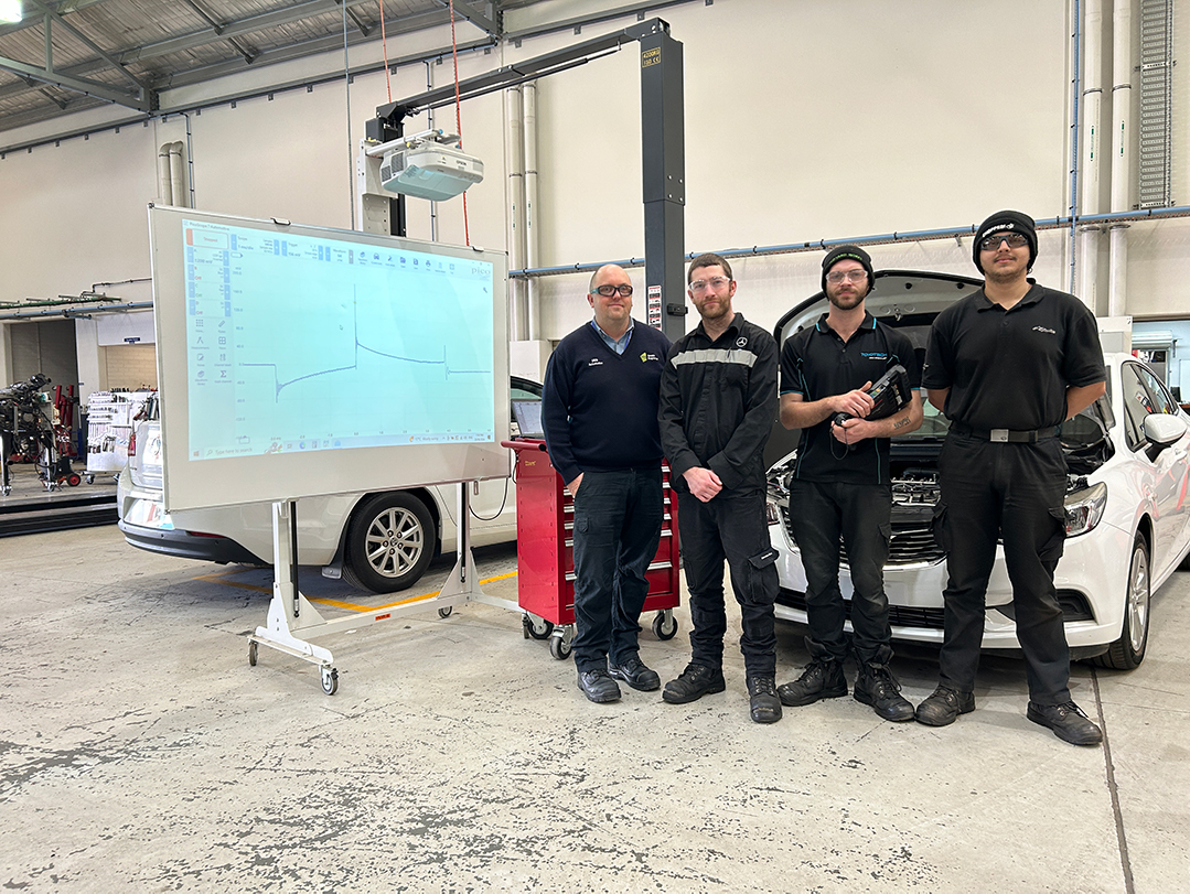 Group of four mechanics standing in an automotive workshop beside a car and a projected diagnostic chart.