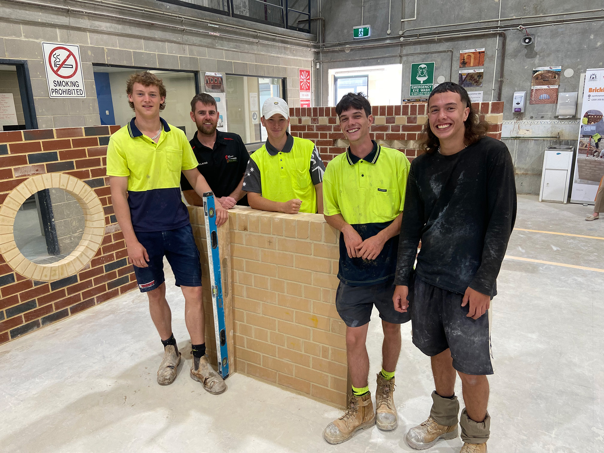  Four young men pose together in front of a textured brick wall, smiling and dressed casually..