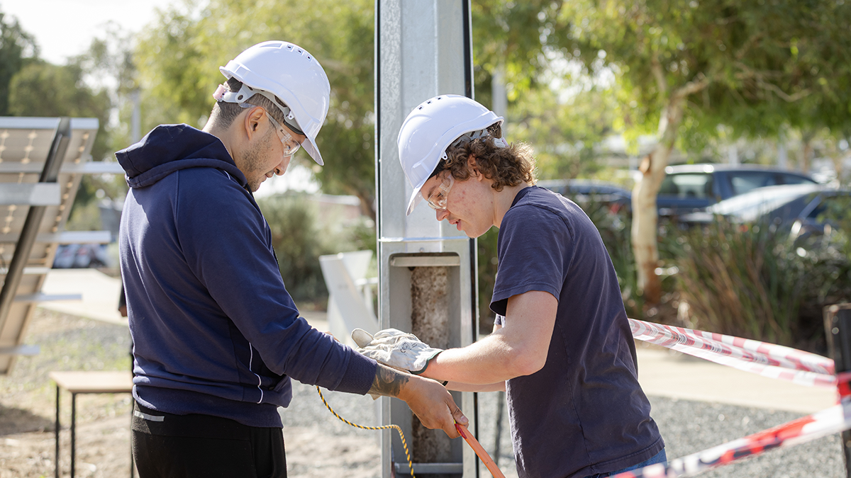 Two people in safety gear working together to adjust a tool on a metal post at an outdoor site, with solar panels and barrier tape in the background.