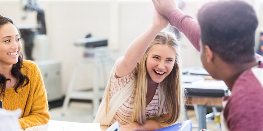 Three teenagers sitting together in a classroom, two are giving each other a high-five.