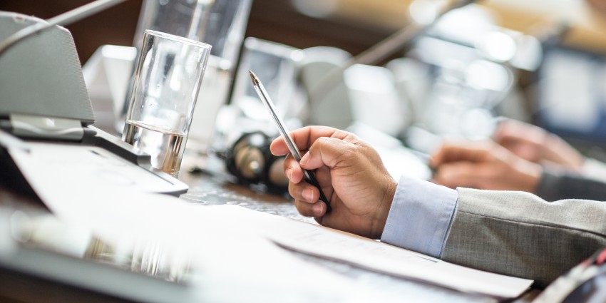 A hand on a desk in an office, holding a pen over some paperwork.