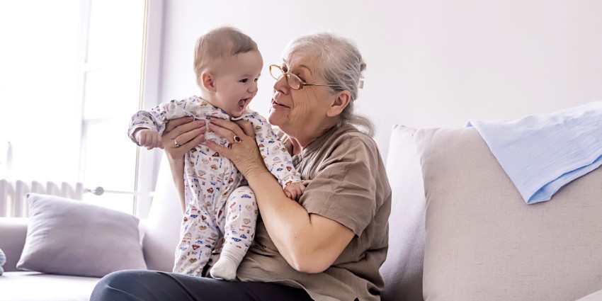 A grandmother holding her small grandchild. They are sitting on a couch, playing.
