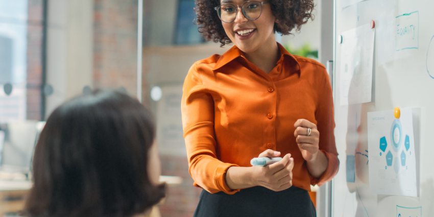 Image of a woman with curly brown hair next to a whiteboard with a pen. She is smiling and speaking to someone in the foreground, another woman with a short bob whose back is to the viewer. 