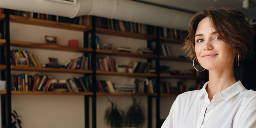 Image of a young woman in a white shirt smiling with arms crossed in the foreground. In the background is an office space with chairs, a laptop on a table and shelves. 