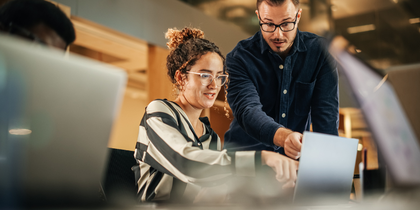 Image of a woman with curly brown hair seated at a laptop with a man wearing glasses and a navy shirt standing beside her. Both of them are pointing at something on the laptop. 