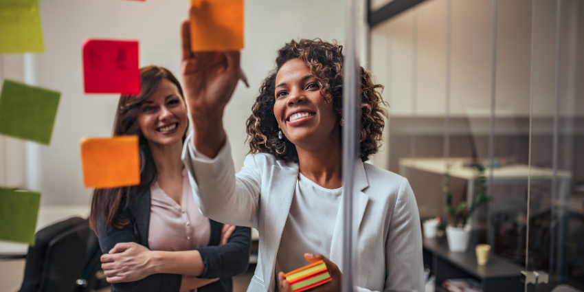 Image of two women standing before a clear glass wall covered with sticky notes. One woman, of African descent in a white top and blazer, is writing something on a sticky note. Another woman is standing behind her, smiling. 