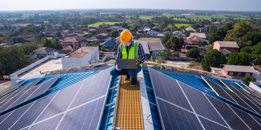 Man crouching on a rooftop surrounded by solar panels. He is holding a laptop.