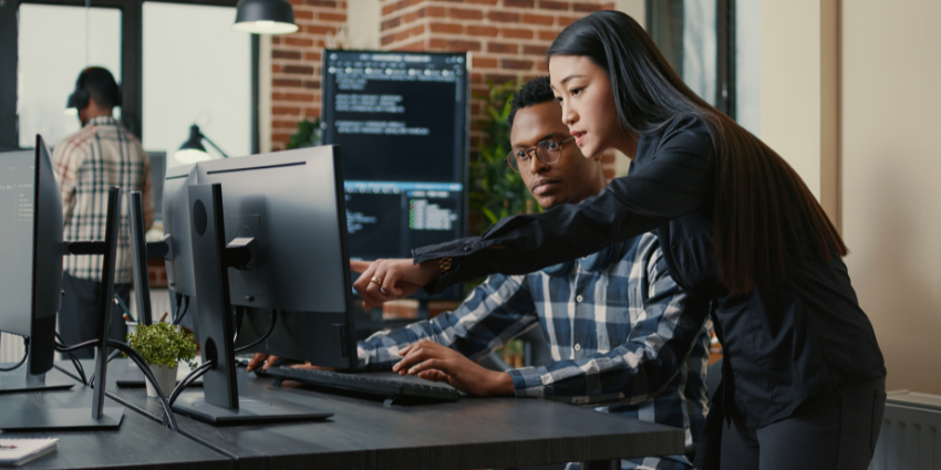 Woman looking over man's shoulder at a desk and pointing at a computer