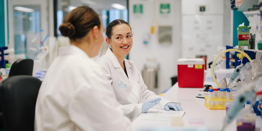 A portrait of Deny Evans wearing a lab coat. She is smiling at a colleague who is in the foreground of the image.