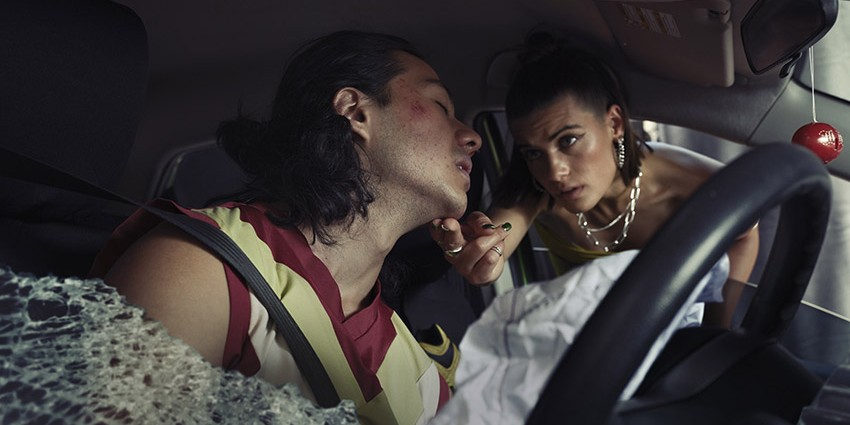 One person is slumped seemingly unconscious in a car after a crash, while another person leans in through the window to check their pulse.
