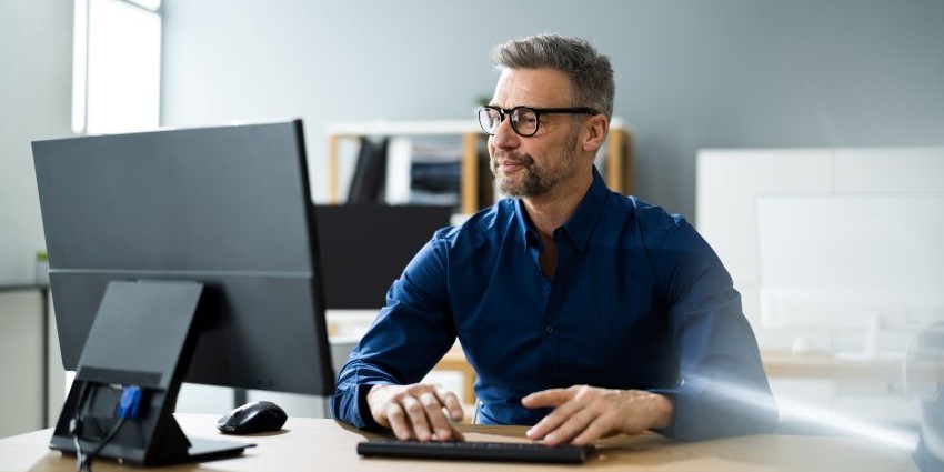 A male office worker with a navy collared shirt and black rimmed glasses looks confidently at his desktop screen,