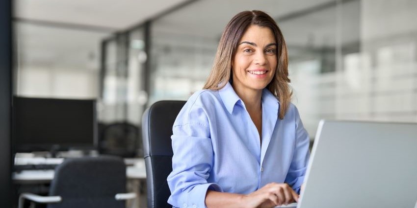 A middle-aged female officer worker smiles at the viewer while sitting at her laptop.