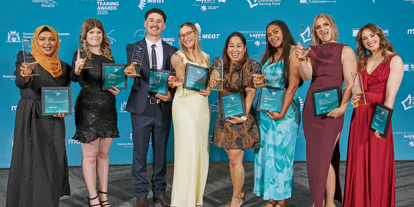 A group of award recipients stand together on a blue event backdrop, each holding a trophy and framed certificate at the WA Training Awards.