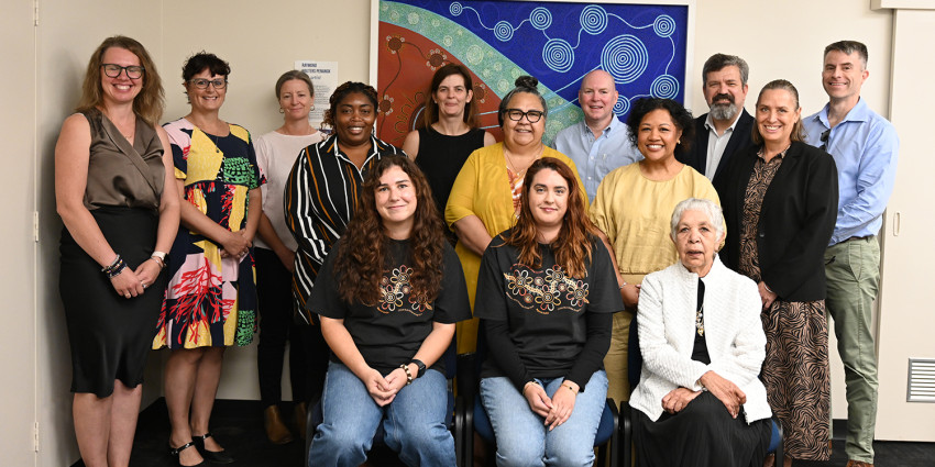 A group shot of people smiling to camera with an Aboriginal painting in the backdrop